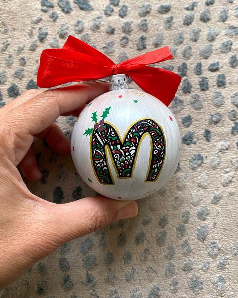 A Pearl white hand painted ball ornament with the letter "M" drawn filled with black doodles and colored with green and red, outlined with gold and green  and red dots scattered all over the ball with gold stars