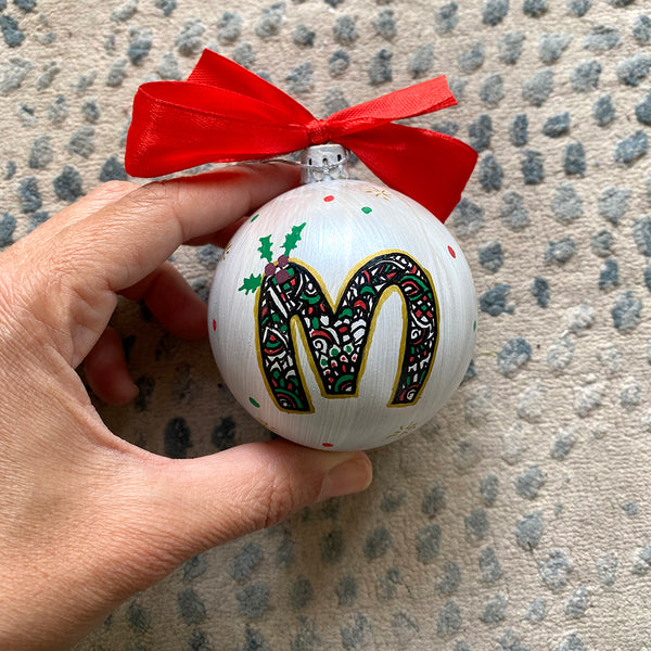 A Pearl white hand painted ball ornament with the letter "M" drawn filled with black doodles and colored with green and red, outlined with gold and green  and red dots scattered all over the ball with gold stars