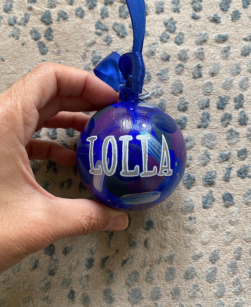 A Colorful hand painted ornament with ultramarine background and colors of ultramarine, dark purple, silver and grey scattered all over with white dots and the name "Lolla" written in silver outlined with White