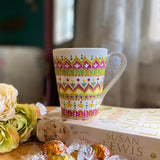 A white mug hand painted with patterns of yellow, olive green and pink, outlined with gold relief and dot art of pink, yellow olive green gold and white