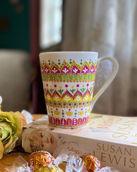 A white mug hand painted with patterns of yellow, olive green and pink, outlined with gold relief and dot art of pink, yellow olive green gold and white