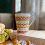 A white mug hand painted with patterns of yellow, olive green and pink, outlined with gold relief and dot art of pink, yellow olive green gold and white