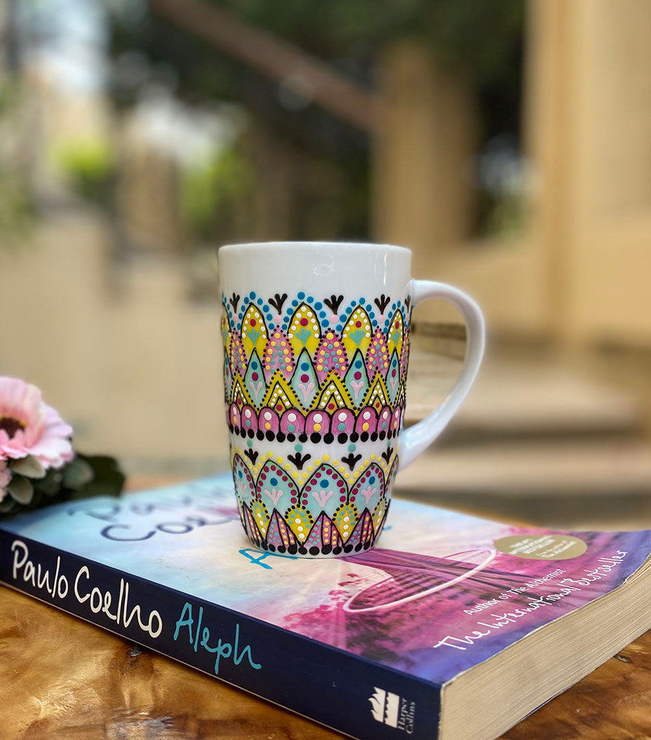 A white mug hand painted with patterns of yellow , pink and mint green , all outlined with black relief relief