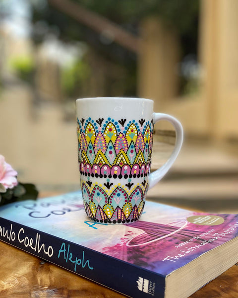 A white mug hand painted with patterns of yellow , pink and mint green , all outlined with black relief relief