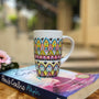 A white mug hand painted with patterns of yellow , pink and mint green , all outlined with black relief relief