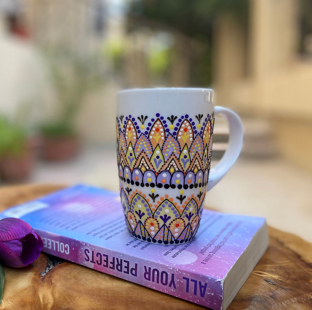 A white mug hand painted with patterns in pastel orange, pastel yellow and lilac, outlined with black relief and dot art of black, purple, lilac, pastel yellow, and pastel orange