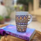 A white mug hand painted with patterns in pastel orange, pastel yellow and lilac, outlined with black relief and dot art of black, purple, lilac, pastel yellow, and pastel orange