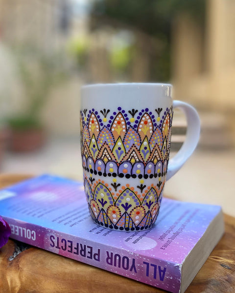 A white mug hand painted with patterns in pastel orange, pastel yellow and lilac, outlined with black relief and dot art of black, purple, lilac, pastel yellow, and pastel orange