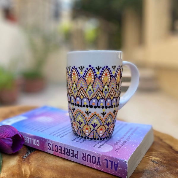 A white mug hand painted with patterns in pastel orange, pastel yellow and lilac, outlined with black relief and dot art of black, purple, lilac, pastel yellow, and pastel orange