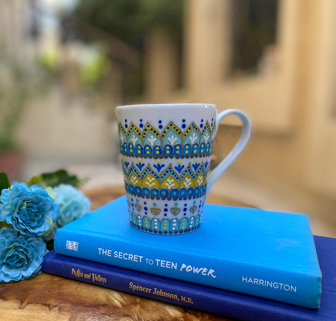 A white mug hand painted with patterns of yellow blue and mint green , all outlined with silver relief