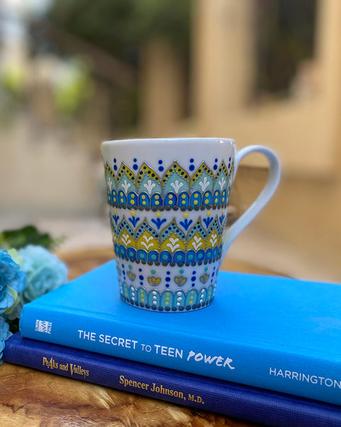 A white mug hand painted with patterns of yellow blue and mint green , all outlined with silver relief