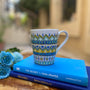 A white mug hand painted with patterns of yellow blue and mint green , all outlined with silver relief