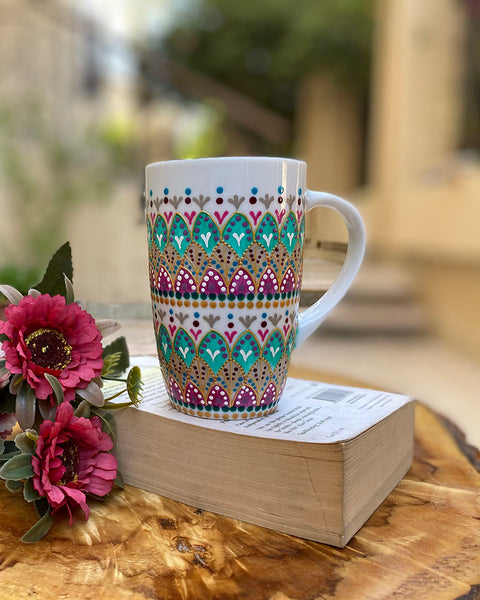 A white mug hand painted with patterns of fushia, taupe and green , all outlined with gold relief