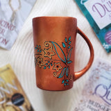 A Copper Hand Painted mug with relief drawings in black and glad of flowers and leaves on curved lines, with leaves colored in blue green