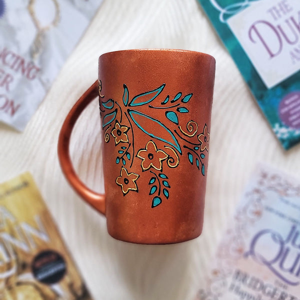 A Copper Hand Painted mug with relief drawings in black and glad of flowers and leaves on curved lines, with leaves colored in blue green