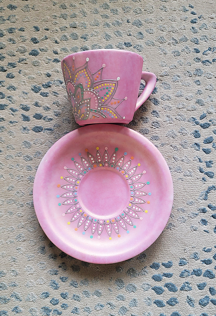 A Shimmery pink hand painted cup and saucer with a mandala drawn in the bottom center spread to both sides using a silver relief and dot art of mint, yellow, baby blue, pastel orange, grey, white and baby pink