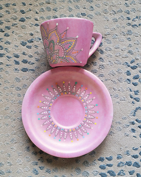 A Shimmery pink hand painted cup and saucer with a mandala drawn in the bottom center spread to both sides using a silver relief and dot art of mint, yellow, baby blue, pastel orange, grey, white and baby pink