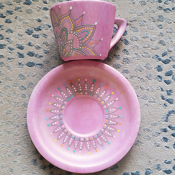 A Shimmery pink hand painted cup and saucer with a mandala drawn in the bottom center spread to both sides using a silver relief and dot art of mint, yellow, baby blue, pastel orange, grey, white and baby pink