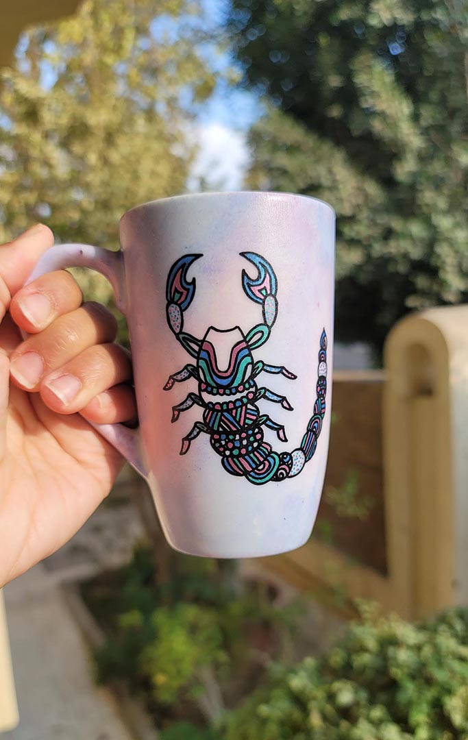 A Pale Hand painted Mug With a Scorpio Design on it in Black and colored in pinks, greens and blues