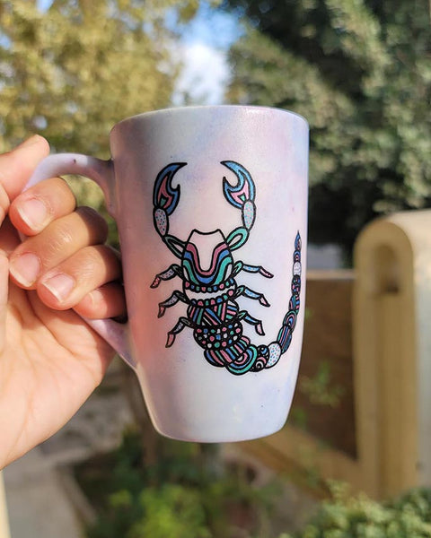 A Pale Hand painted Mug With a Scorpio Design on it in Black and colored in pinks, greens and blues