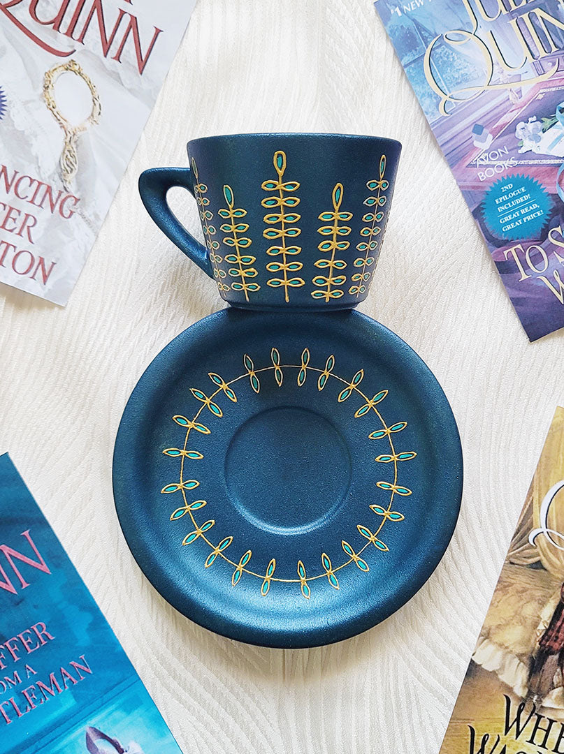 A blue shimmery hand painted cup and saucer with relief vertical leaves drawn in gold all around the cup and all around the plate. Colored in with blue green outlines