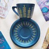 A blue shimmery hand painted cup and saucer with relief vertical leaves drawn in gold all around the cup and all around the plate. Colored in with blue green outlines