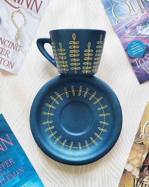 A blue shimmery hand painted cup and saucer with relief vertical leaves drawn in gold all around the cup and all around the plate. Colored in with blue green outlines