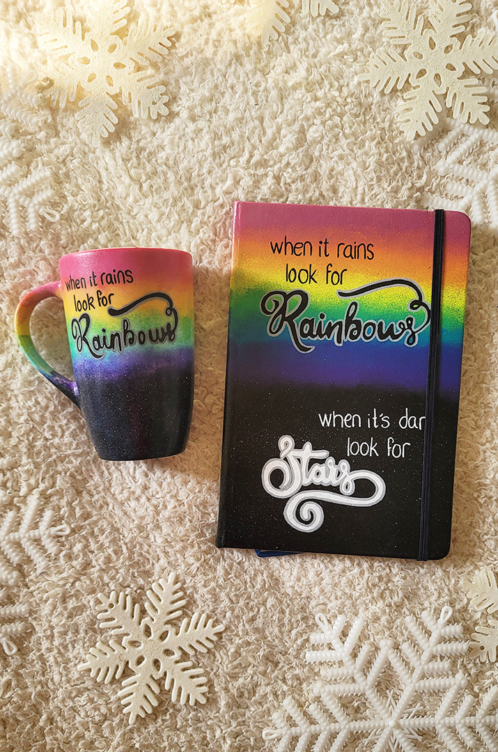 A hand painted Mug and notebook with the top half painted in rainbow and the bottom in dark galaxy, with the words on the rainbow written "when it rains look for rainbows"