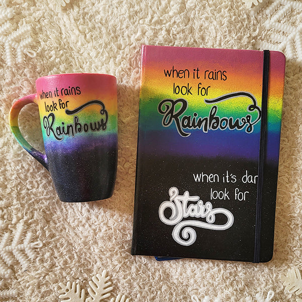 A hand painted Mug and notebook with the top half painted in rainbow and the bottom in dark galaxy, with the words on the rainbow written "when it rains look for rainbows"