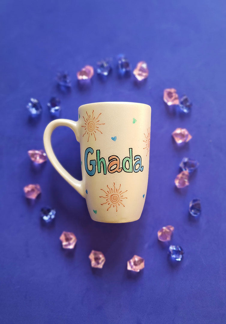 An Ivory Hand painted mug with the name ghada and small doodle suns and small hearts scattered all over the mug