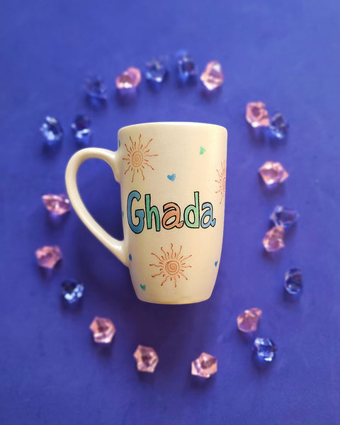 An Ivory Hand painted mug with the name ghada and small doodle suns and small hearts scattered all over the mug