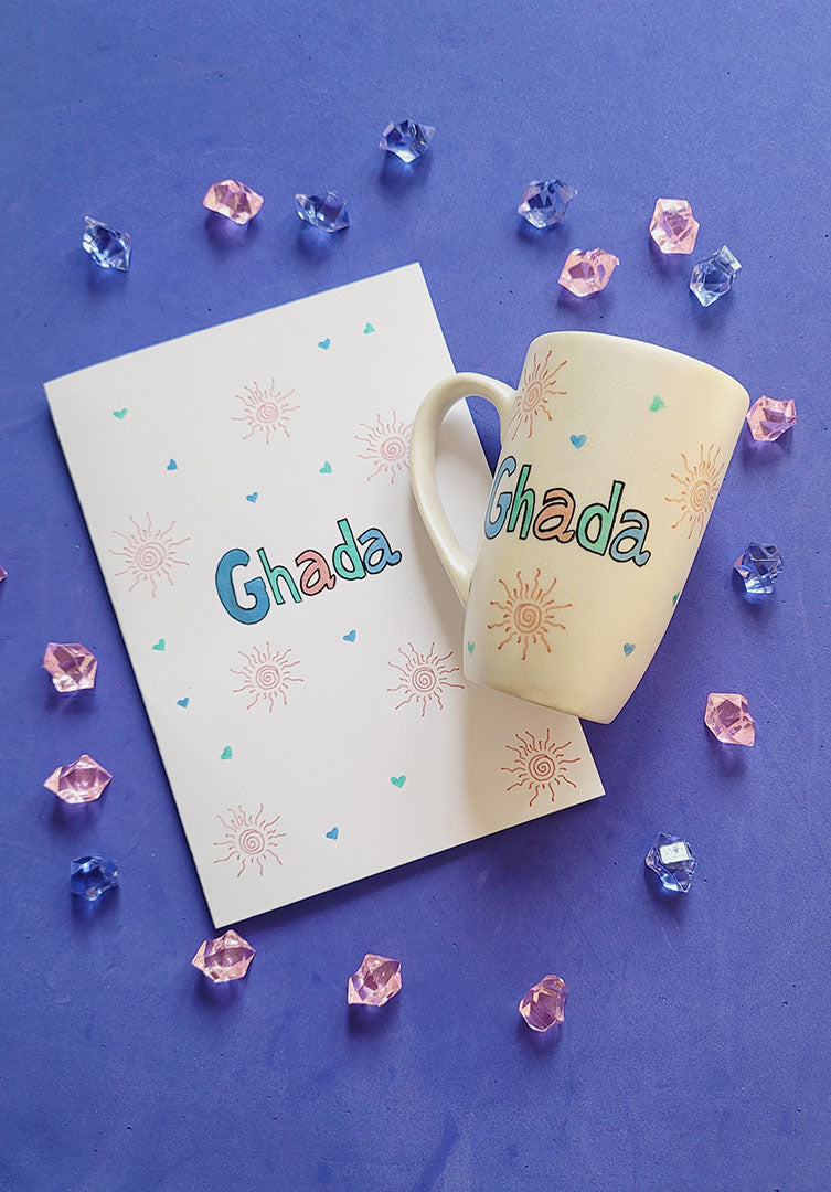 An Ivory Hand painted mug and Card with the name ghada and small doodle suns and small hearts scattered all.