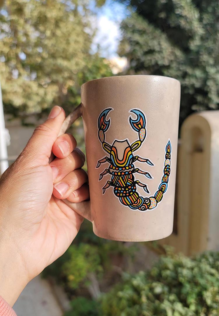 A Taupe Hand painted Mug With a Scorpio Design on it in Black and colored in yellow, orange and blue