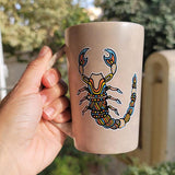 A Taupe Hand painted Mug With a Scorpio Design on it in Black and colored in yellow, orange and blue