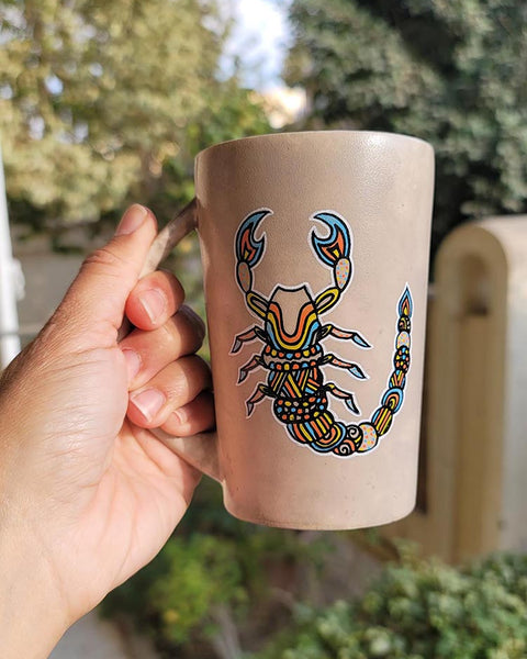 A Taupe Hand painted Mug With a Scorpio Design on it in Black and colored in yellow, orange and blue