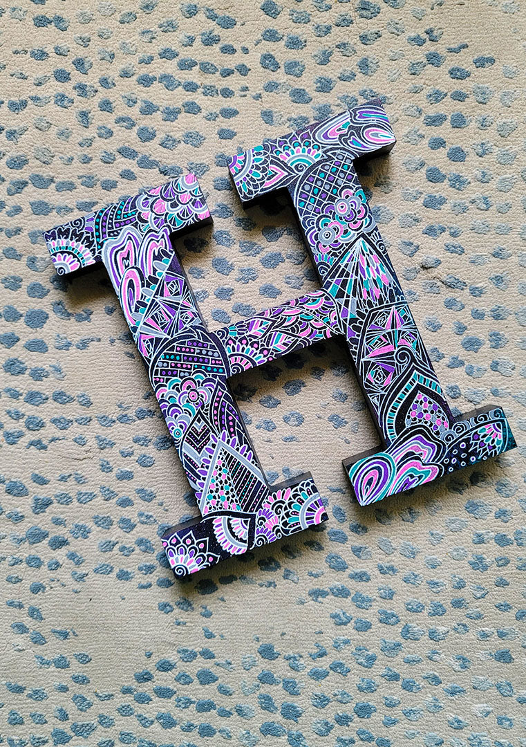 A black galaxy hand painted letter H with doodles in white and colored in with pink , turquoise, purple and grey 