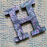 A black galaxy hand painted letter H with doodles in white and colored in with pink , turquoise, purple and grey 