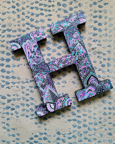 A black galaxy hand painted letter H with doodles in white and colored in with pink , turquoise, purple and grey 