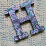 A black galaxy hand painted letter H with doodles in white and colored in with pink , turquoise, purple and grey 