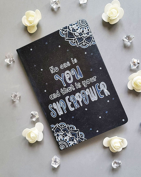 A Black Galaxy Hand painted notebook with 2 small mandala drawn in white in the opposite corners and the quote written in the center "no one is You and that is your Superpower"