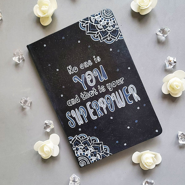 A Black Galaxy Hand painted notebook with 2 small mandala drawn in white in the opposite corners and the quote written in the center "no one is You and that is your Superpower"