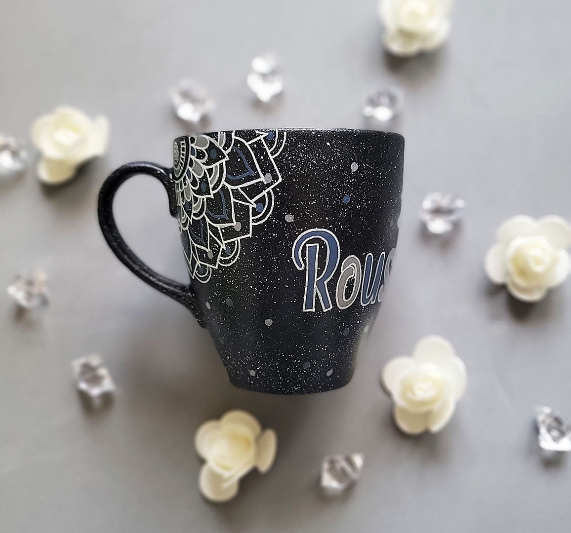 A bLack Galaxy hand painted mug with the name rousha in the middle and ont he corners a mandala drawn in white with patterns on dark grey and light grey. And light grey and dark grey dots scattered all over the mug