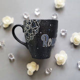 A bLack Galaxy hand painted mug with the name rousha in the middle and ont he corners a mandala drawn in white with patterns on dark grey and light grey. And light grey and dark grey dots scattered all over the mug