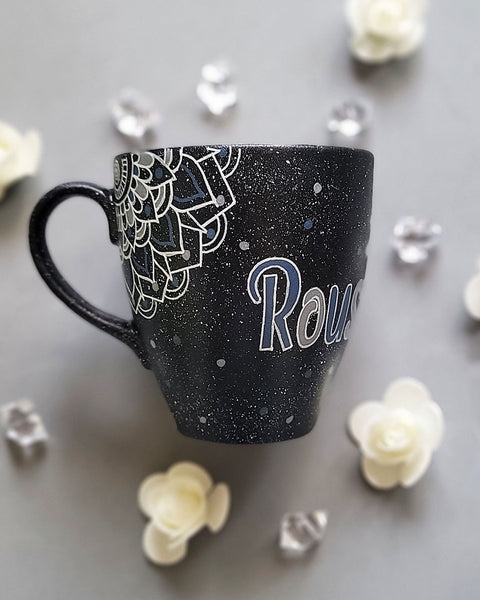 A bLack Galaxy hand painted mug with the name rousha in the middle and ont he corners a mandala drawn in white with patterns on dark grey and light grey. And light grey and dark grey dots scattered all over the mug