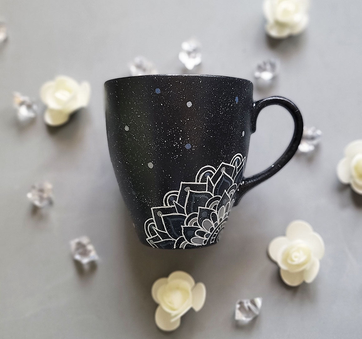 A bLack Galaxy hand painted mug with the name rousha in the middle and ont he corners a mandala drawn in white with patterns on dark grey and light grey. And light grey and dark grey dots scattered all over the mug