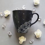 A bLack Galaxy hand painted mug with the name rousha in the middle and ont he corners a mandala drawn in white with patterns on dark grey and light grey. And light grey and dark grey dots scattered all over the mug