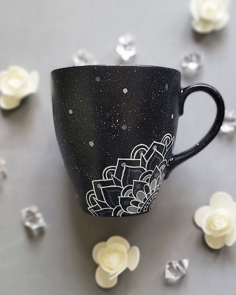 A bLack Galaxy hand painted mug with the name rousha in the middle and ont he corners a mandala drawn in white with patterns on dark grey and light grey. And light grey and dark grey dots scattered all over the mug