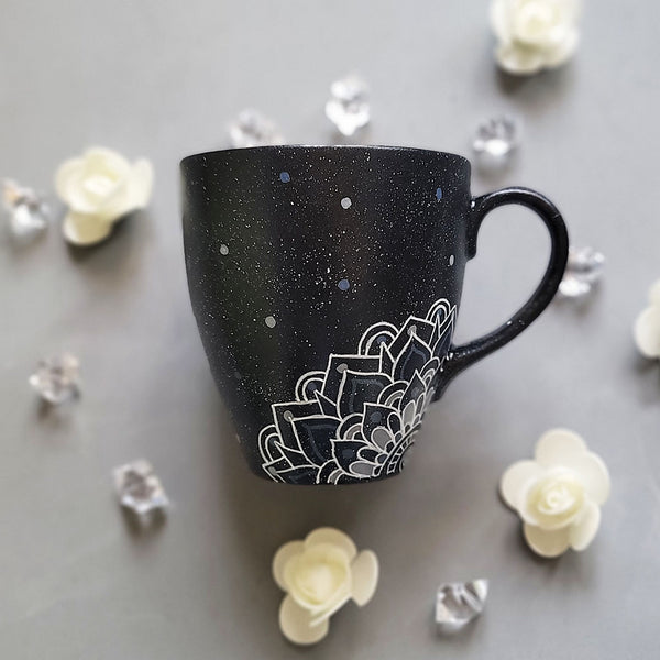 A bLack Galaxy hand painted mug with the name rousha in the middle and ont he corners a mandala drawn in white with patterns on dark grey and light grey. And light grey and dark grey dots scattered all over the mug