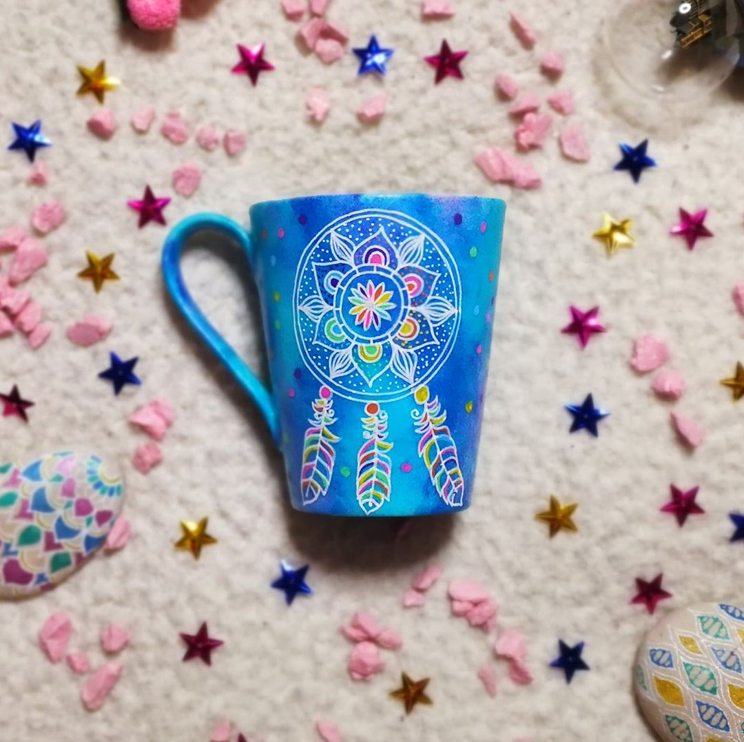 A Blue hand painted mug with a dreamcatcher in white and colored in with (pink, yellow, purple blue, green and orange)