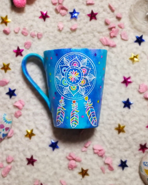 A Blue hand painted mug with a dreamcatcher in white and colored in with (pink, yellow, purple blue, green and orange)